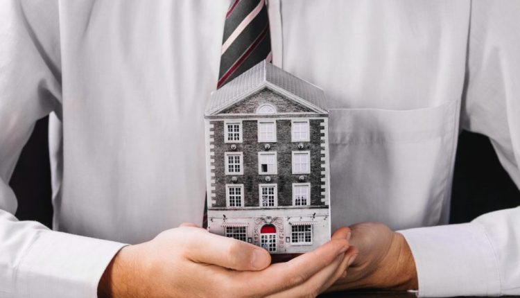 Person in a white shirt and striped tie holds a small brick house model on a wooden table