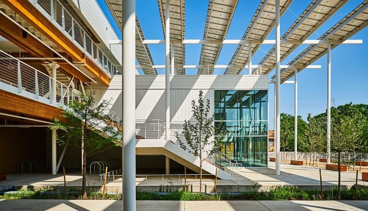 Modern building exterior with glass facade, white metal beams, and a covered walkway; trees and blue sky visible.
