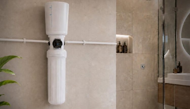 Vertical wall-mounted water filtration unit in a beige tiled bathroom, with white cylindrical filter housing and exposed piping nearby.